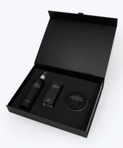 A tall cylindrical matte plastic bottle with a clear plastic lid that has London Grooming Company Sea Salt Spray written in bright white writing on it next to a short rectangular cardboard box that has London Grooming Company Volumizing Dust written in bright white writing on it, and a short cylindrical black tin that has London Grooming Company Hair Clay written in bright white writing on it inside a large rectangular black cardboard box, on a white background.