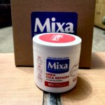 A Mixture Of Urea & Cica Is Great For Dry & Rough Skin | Mixa Urea Cica Repair+ Renewing Cream | Gifted A tall cylindrical bright white plastic tub with a bright white plastic lid and a bright red label that has Mixa written in bright white writing inside a dark blue rectangle and Urea Cica Repair+ Renewing Cream written in bright white writing inside a bright red rectangle on it in front of a large rectangular cardboard box that has Mixa written in dark blue writing on it on a light wooden table, on a white background.