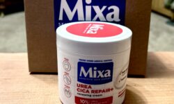 A tall cylindrical bright white plastic tub with a bright white plastic lid and a bright red label that has Mixa written in bright white writing inside a dark blue rectangle and Urea Cica Repair+ Renewing Cream written in bright white writing inside a bright red rectangle on it in front of a large rectangular cardboard box that has Mixa written in dark blue writing on it on a light wooden table, on a white background.