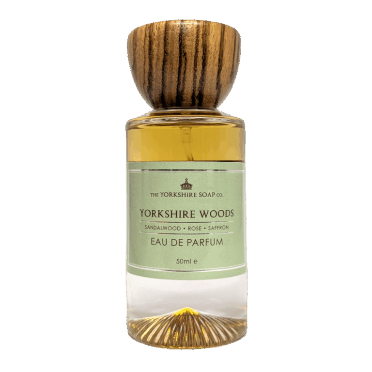 A tall cylindrical clear glass bottle full of some golden amber liquid with a circular dark wooden lid and a pale green label that has The Yorkshire Soap Company Yorkshire Woods Eau De Parfum written in bold black writing and a picture of a black crown on it, on a white background.