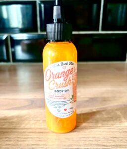 A short cylindrical clear plastic bottle full of some shimmering bright orange liquid with a black plastic nozzle lid and a bright white label that has Orange Crush written in bright orange writing and Bath Bomb Bliss Body Oil written in bold black writing on it, on a light wooden worktop.