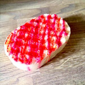A large creamy white oval Sponge that has some grooves and ridges all over it and some bright red soap engraved on it, on a light wooden worktop.