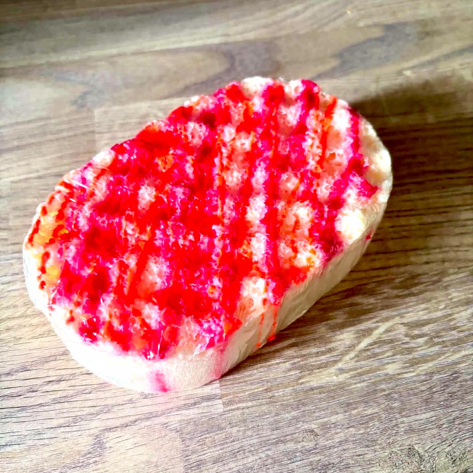 A large creamy white oval Sponge that has some grooves and ridges all over it and some bright red soap engraved on it, on a light wooden worktop.