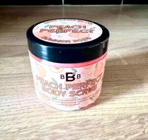 A short cylindrical bright white plastic tub with a black plastic lid and a bright peach label that has Bath Bomb Bliss written in dark red writing, Peach Perfect Body Scrub written in bright pink writing and some pictures of some bright pink whole and sliced peaches on it, on a light wooden worktop.