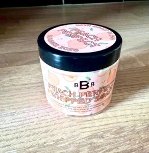 A short cylindrical bright white plastic tub with a black plastic lid and a bright peach label that has Bath Bomb Bliss written in dark red writing, Peach Perfect Whipped Soap written in bright pink writing and some pictures of some bright pink whole and sliced peaches on it, on a light wooden worktop.
