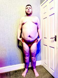 A tall bearded man with his arms resting by his sides wearing a bright orange thong with a bright orange waistband that has Kiniki written in bright white writing on it stood against a white wooden door.