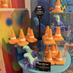 Several bright orange Traffic cone shaped Bath Bombs that have a bright white stripe around the centre of them inside some medium clear acrylic boxes on a bright turquoise shelf next to several bright green loch ness monster shaped bath bombs that have some bright green heads, necks and upper bodies and a dark purple underneath on them, on a white background.