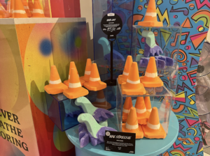 Several bright orange Traffic cone shaped Bath Bombs that have a bright white stripe around the centre of them inside some medium clear acrylic boxes on a bright turquoise shelf next to several bright green loch ness monster shaped bath bombs that have some bright green heads, necks and upper bodies and a dark purple underneath on them, on a white background.