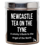 Tea Tastes Terrific | Bird & Blend Tea Company Newcastle Tea On The Tyne Loose Leaf Tea A tall cylindrical bright silver metal tin with a bright silver metal lid and a black label that has Newcastle Tea On The Tyne Loose Leaf Tea, a smoky tribute to the Angel of the North written in bright white writing and a picture of a plant with two leaves and a flower bud attached to it on it, on a white background.