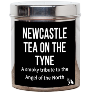 A tall cylindrical bright silver metal tin with a bright silver metal lid and a black label that has Newcastle Tea On The Tyne Loose Leaf Tea, a smoky tribute to the Angel of the North written in bright white writing and a picture of a plant with two leaves and a flower bud attached to it on it, on a white background.