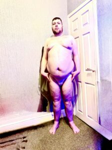 A tall bearded man with his hands on his outer thighs wearing a pair of light beige briefs with a black waistband that has Box Menswear written in bright white writing on it stood against a white wooden door.