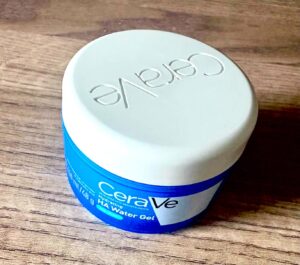 A short cylindrical bright blue plastic tub with a bright white plastic lid and a bright white label that has Cera written in bright white writing, Ve written in bright blue writing inside a slanted bright white rectangle, Hydrating HA Water Gel written in bright white writing inside a bright blue rectangle, and CeraVe engraved on it, on a light wooden worktop.