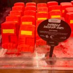 London Calling To The Faraway Towns | Lush Double Decker Bus Bath Bomb Several medium rectangular bright red Double Decker bus shaped bath bombs that have a bright yellow panel that has London written in bright yellow writing on it surrounded by some dark red bath bomb dust inside a light wooden tray next to a round black cardboard sign that has Oxford Street Exclusive and Double Decker Bus Bath Bomb written in bold black writing inside a bright white rectangle on it, on a white background.