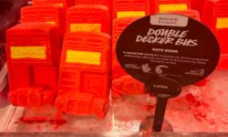 Several medium rectangular bright red Double Decker bus shaped bath bombs that have a bright yellow panel that has London written in bright yellow writing on it surrounded by some dark red bath bomb dust inside a light wooden tray next to a round black cardboard sign that has Oxford Street Exclusive and Double Decker Bus Bath Bomb written in bold black writing inside a bright white rectangle on it, on a white background.