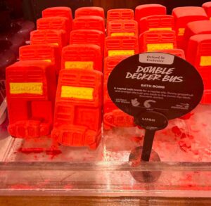 Several medium rectangular bright red Double Decker bus shaped bath bombs that have a bright yellow panel that has London written in bright yellow writing on it surrounded by some dark red bath bomb dust inside a light wooden tray next to a round black cardboard sign that has Oxford Street Exclusive and Double Decker Bus Bath Bomb written in bold black writing inside a bright white rectangle on it, on a white background.
