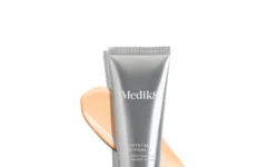 A tall conical metallic silver plastic tube with a metallic silver pump lid that has Medik8 Crystal Retinal written in bright white writing on it surrounded by some large streaks of some creamy beige Facial Serum, on a white background.