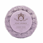 Soaking Up Yorkshire's Culture Like A Big Sponge | The Yorkshire Soap Company Soap Sponge Range A large creamy lilac circular Sponge that has some grooves and ridges all over it and some clear soap engraved on it with a circular pale lilac label that has The Yorkshire Soap Company Cherry Noir Soap Sponge written in dark lilac writing and a picture of a dark lilac crown on it wrapped in some clear plastic, on a white background.