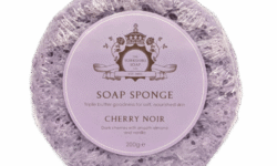 A large creamy lilac circular Sponge that has some grooves and ridges all over it and some clear soap engraved on it with a circular pale lilac label that has The Yorkshire Soap Company Cherry Noir Soap Sponge written in dark lilac writing and a picture of a dark lilac crown on it wrapped in some clear plastic, on a white background.