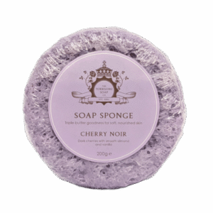 A large creamy lilac circular Sponge that has some grooves and ridges all over it and some clear soap engraved on it with a circular pale lilac label that has The Yorkshire Soap Company Cherry Noir Soap Sponge written in dark lilac writing and a picture of a dark lilac crown on it wrapped in some clear plastic, on a white background.