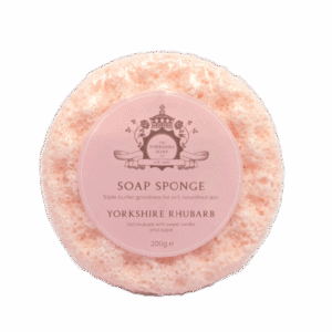 A large creamy pink circular Sponge that has some grooves and ridges all over it and some clear soap engraved on it with a circular pale pink label that has The Yorkshire Soap Company Yorkshire Rhubarb Soap Sponge written in dark pink writing and a picture of a dark pink crown on it wrapped in some clear plastic, on a white background.