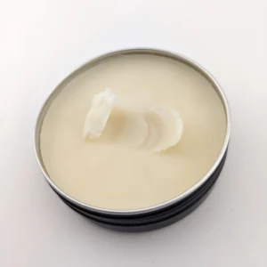 A short cylindrical shiny Black metal tin full of some pale creamy beard butter that has a silver metal inside, on a white background.
