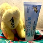The Mother Of Soothed & Repaired skin | BioGaia Aldermis Baby Ointment | Gifted A short slightly tapered light blue plastic tube with a bright white Plastic lid that has BioGaia written in dark blue writing and a picture of a bright yellow baby on it next to a small bright yellow Flying baby shaped plushie with some bright yellow Arms, some bright yellow legs and a bright yellow cape that has BioGaia written in bold black writing on it on top of a piece of bright teal green fabric that has some abstract bright red stylised letters all over it, on a light wooden table.