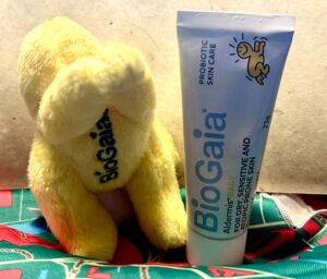 A short slightly tapered light blue plastic tube with a bright white Plastic lid that has BioGaia written in dark blue writing and a picture of a bright yellow baby on it next to a small bright yellow Flying baby shaped plushie with some bright yellow Arms, some bright yellow legs and a bright yellow cape that has BioGaia written in bold black writing on it on top of a piece of bright teal green fabric that has some abstract bright red stylised letters all over it, on a light wooden table.