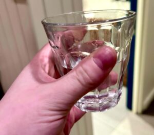 A hand holding a short cylindrical faceted glass tumbler full of a very little amount of clear water, on a white background.