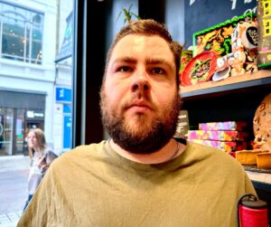 A tall bearded man wearing a bright gold chain and a dark Olive Green crew neck t-shirt stood in front of a large rectangular shelving unit full of several large rectangular multicoloured boxes and some different multicoloured bath bombs next to a large rectangular window looking on to the Street, on a white background.