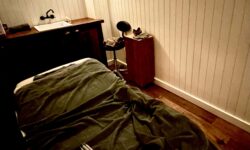 A large rectangular spa treatment bed that’s covered with a dark green loosely draped and wrinkled Blanket next to a medium rectangular light wooden padded table, a dark wooden counter that has a bright white porcelane sink with a black tap embedded in it and a folded up dark green towel on it, a small tall rectangular dark wooden table that has a folded up dark green towel and a pair of dark red shoes on top of it, and a medium black rounded stool with some metal legs inside a large room that has some creamy white vertical panelling and a dark wooden floor, on a white background.