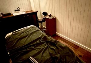 A large rectangular spa treatment bed that’s covered with a dark green loosely draped and wrinkled Blanket next to a medium rectangular light wooden padded table, a dark wooden counter that has a bright white porcelane sink with a black tap embedded in it and a folded up dark green towel on it, a small tall rectangular dark wooden table that has a folded up dark green towel and a pair of dark red shoes on top of it, and a medium black rounded stool with some metal legs inside a large room that has some creamy white vertical panelling and a dark wooden floor, on a white background.