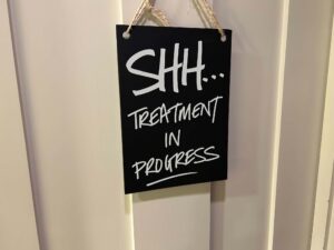 A large rectangular light wooden panelled door with a rectangular black sign that has Shh…! Treatment In Progress written in bright white writing on it, on a white background.