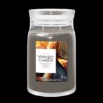 The Last Glow Of Summer Means That Autumn Has Finally Arrived | Yankee Candle Glowing Moments Collection A tall cylindrical glass Jar full of some pale Grey Wax with a bright white label that has Yankee Candle written in bold black writing, Crackling Embers written in bold black writing, and a picture of some burning kindling and some bright red, orange and yellow glowing embers on it, on a black background.