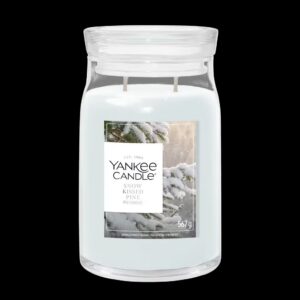A tall cylindrical glass Jar full of some creamy white Wax with a bright white label that has Yankee Candle written in bold black writing, Snow Kissed Pine written in bold black writing, and a picture of some dark green Pine Branches covered in some bright white Snow on it, on a black background.