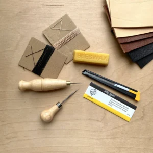 Several small rectangular pieces of light tan, brown and black leather next to a piece of black thread wrapped around a small rectangular piece of cardboard, a piece of tan thread wrapped around a small rectangular piece of cardboard, a large rectangular block of yellow beeswax that has Beeswax engraved on it, a medium black and yellow snap-off utility knife, a long and thin black and yellow paper packet of needles, and two wooden handle Awls, on a light wooden table.