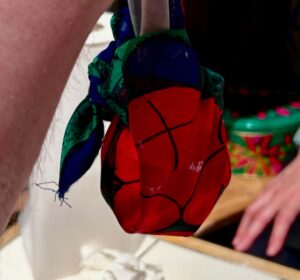 A medium circular bath bomb wrapped in a piece of glossy bright red cloth that has some thin black petal drawings and some dark blue and green leaf drawings all over it with a knot at the top of it on a creamy white paper covered table, on a white background.
