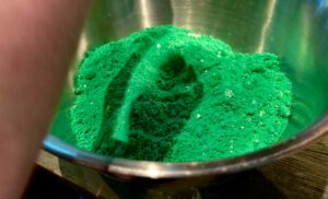 A large cylindrical metal mixing bowl full of some bright green Bath Bomb Powder that has some specks of dark Green Bath Bomb Powder running through it on a light wooden table, on a white background.