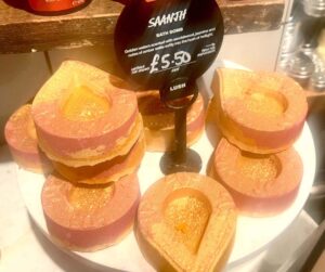A pile of several gradient bright peach and red flame shaped bath bombs that all have some bright gold lustre all over them on a bright white circular plate next too a large black circular sign that has Lush Saanjh Bath Bomb written in bright white writing on it on a light wooden table, on a white background.