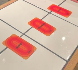 A small rectangular Tarot Card that has Lush Energy written in bold black writing and a picture of a bright Orange and red concentric circle pattern on it next to a small rectangular Tarot Card that has Lush Confidence written in bold black writing and a picture of a bright Orange and red concentric circle pattern on it, and a small rectangular Tarot Card that has Lush Comfort written in bold black writing and a picture of a bright Orange and red concentric circle pattern on it on a light wooden table that’s covered in some bright white tiles, on a white background.