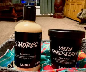 A tall cylindrical clear plastic bottle full of some creamy beige shower gel with a black label that has Lush S’mores Shower Gel written in bright white writing on it next to a tall cylindrical black plastic tub with a black label that has Lush Yuzu Cheesecake Body Butter written in bright white writing on it inside a bright red, orange and turquoise fabric lined light grey fabric basket on a light wooden table, on a white background.
