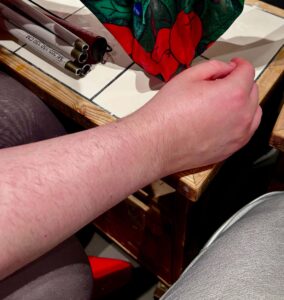 A light-skinned forearm with it’s hand turned slightly inward resting on a light wooden table that’s covered in some matte white Tiles, on a white background.