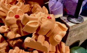 A pile of several large bright orange reindeer shaped bath bombs that all have some red noses, some big ears, some round snouts, and some raised eyes, legs and antlers engraved on them inside a large rectangular wooden box, on a white background.