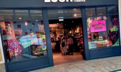 A large rectangular brightly lit glass store front with a large rectangular black board over it’s door that has Lush Fresh Handmade Cosmetics written in bright white writing on it and a picture of a rainbow on one of it’s windows, on a white background.