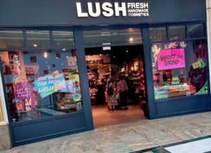 A large rectangular brightly lit glass store front with a large rectangular black board over it’s door that has Lush Fresh Handmade Cosmetics written in bright white writing on it and a picture of a rainbow on one of it’s windows, on a white background.