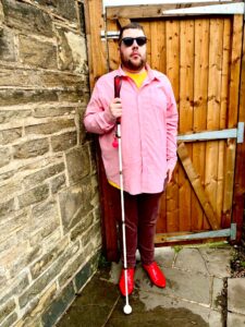 A tall bearded man wearing a pair of dark denim blue glasses with some pale red lenses, a bright pink long sleeve Oxford Shirt, a golden orange crew neck t-shirt, a pair of dark burgundy chino trousers and a pair of shiny bright red shoes holding a long bright white cane with a bright red leather handle stood in front of a medium brown gate.